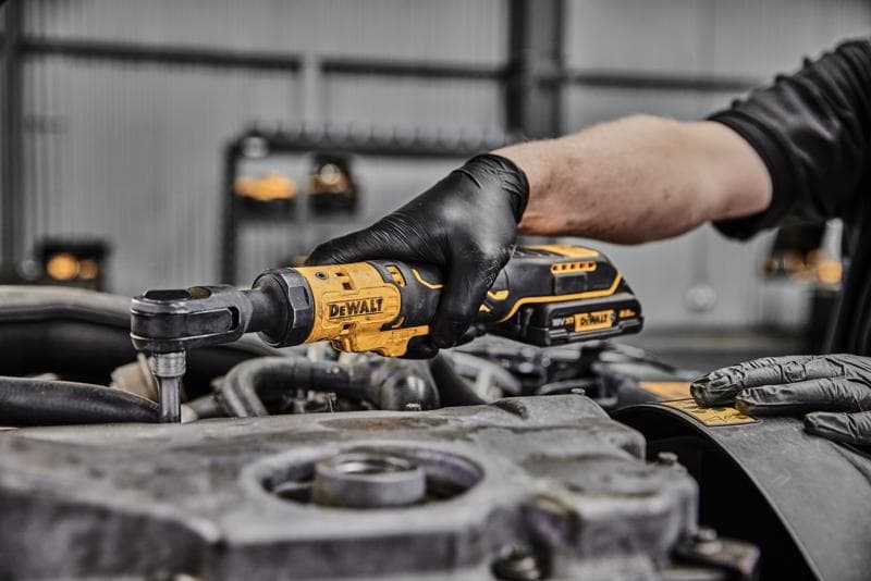 A person wearing black gloves uses a DEWALT cordless ratchet tool on an engine component in a workshop.