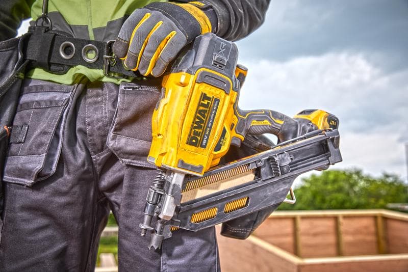 Close-up of a person wearing work gloves holding a DEWALT DCN930N nailer at a construction site, with a wooden structure in the background.