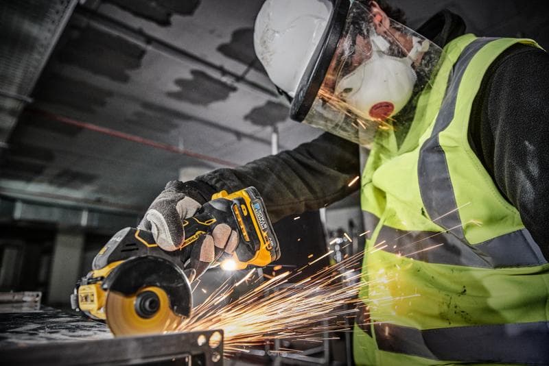 A worker wearing protective gear, including a hard hat, face shield, gloves, and a high-visibility vest, uses a DEWALT cordless angle grinder (model DCS438N) to cut metal, producing sparks.