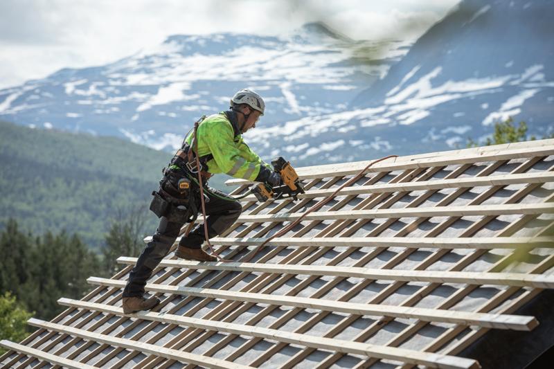 A construction worker wearing safety gear uses a DEWALT cordless nailer to attach wooden battens to a sloped roof, with a scenic mountain landscape in the background.