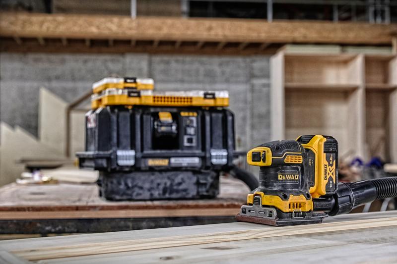 A DEWALT cordless sander (DCW200P2) is placed on a wooden plank, with a DEWALT tool storage box in the background inside a workshop setting. The sander is connected to a hose for dust extraction.
