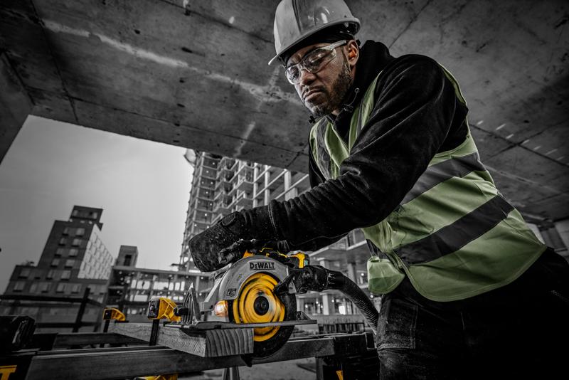 A construction worker wearing a safety vest and helmet operates a DEWALT circular saw (model DCS565NT) to cut a wooden plank at a building site. The background shows concrete structures and high-rise buildings under construction.